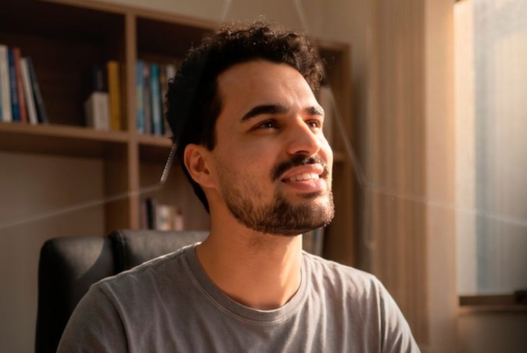 Lucas de Oliveira sorrindo, sentado em um escritório com estante de livros